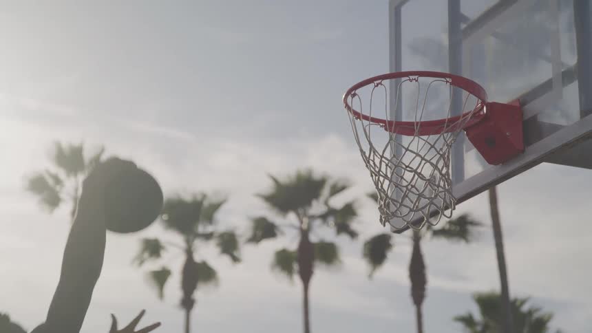 In a hazy sunlit outdoor basketball court an arm reaches high to shoot a basketball poised against the backdrop of palm trees