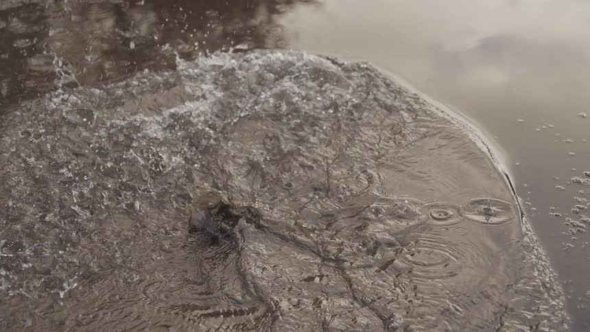A brown leather boot splashes into a muddy puddle creating a large splash and ripples in the water reflecting the sky