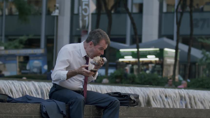 Man in shirt and tie sitting next to a fountain on a sidewalk by a fountain in new york city eating a snack