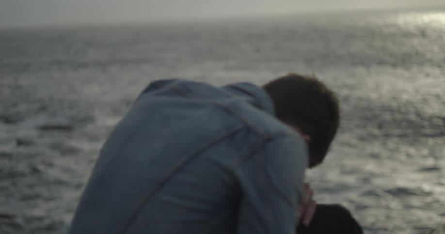 Man with his head down near the water on a cloudy day
