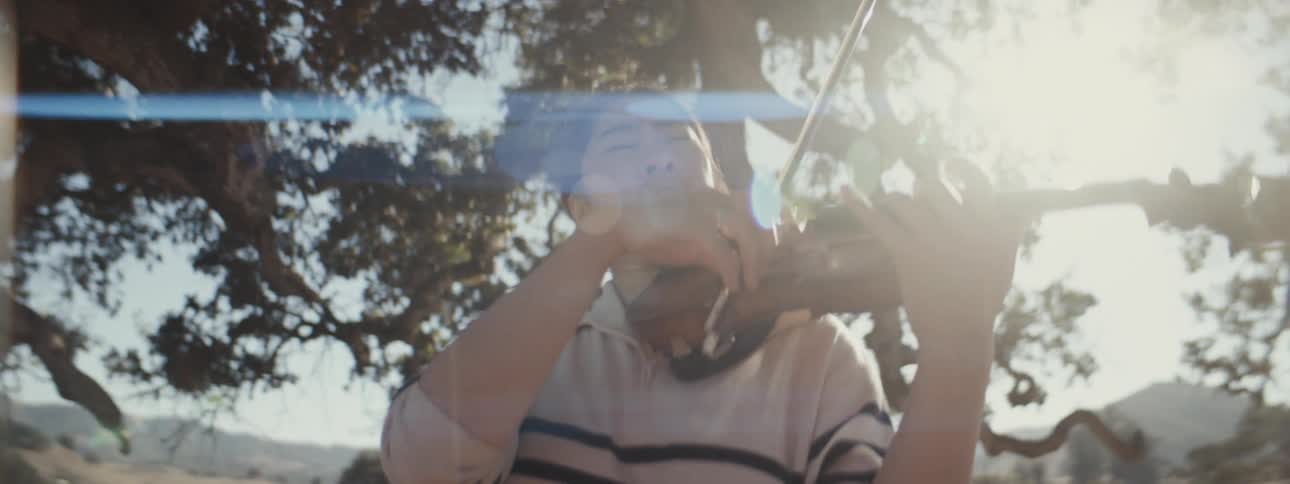 A person is playing a violin outdoors under a large tree with sunlight filtering through the branches wearing a striped longsleeve shirt