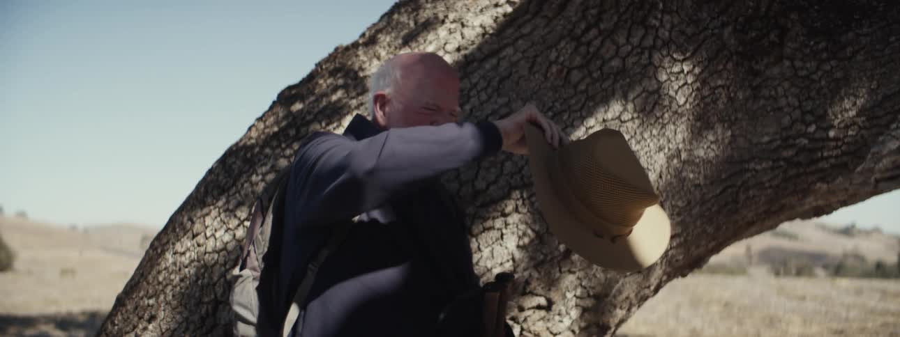 Elderly man walking outdoors and stopping next to a tree trunk and wiping sweat from his head