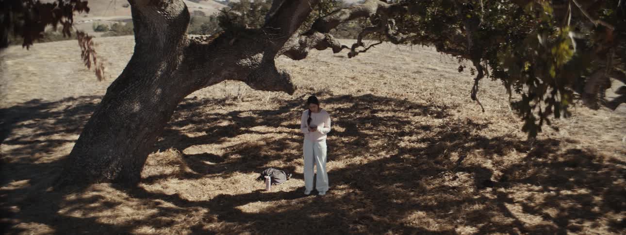 A woman in white clothes stands next to a large tree and then starts walking and sits on the ground