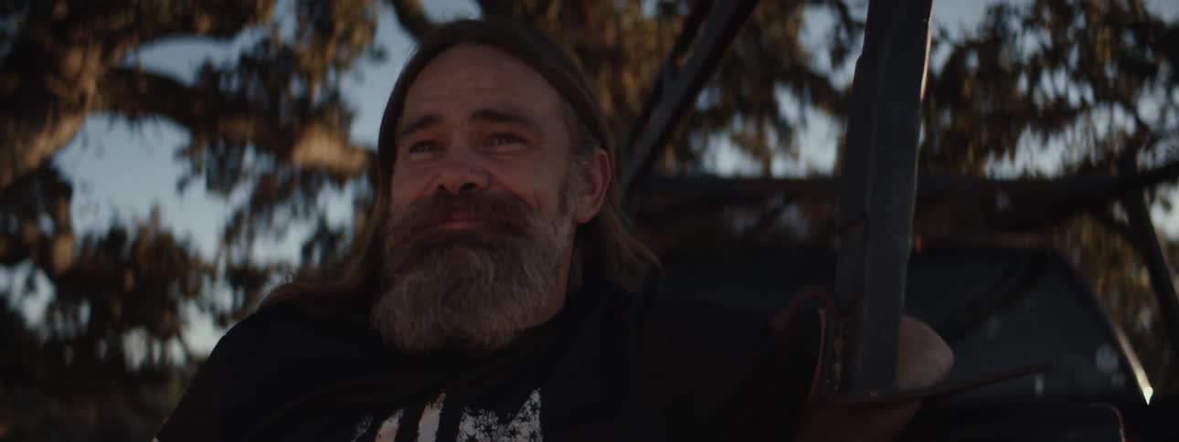 The face of a bearded man drinking a beer outdoors while talking to the front