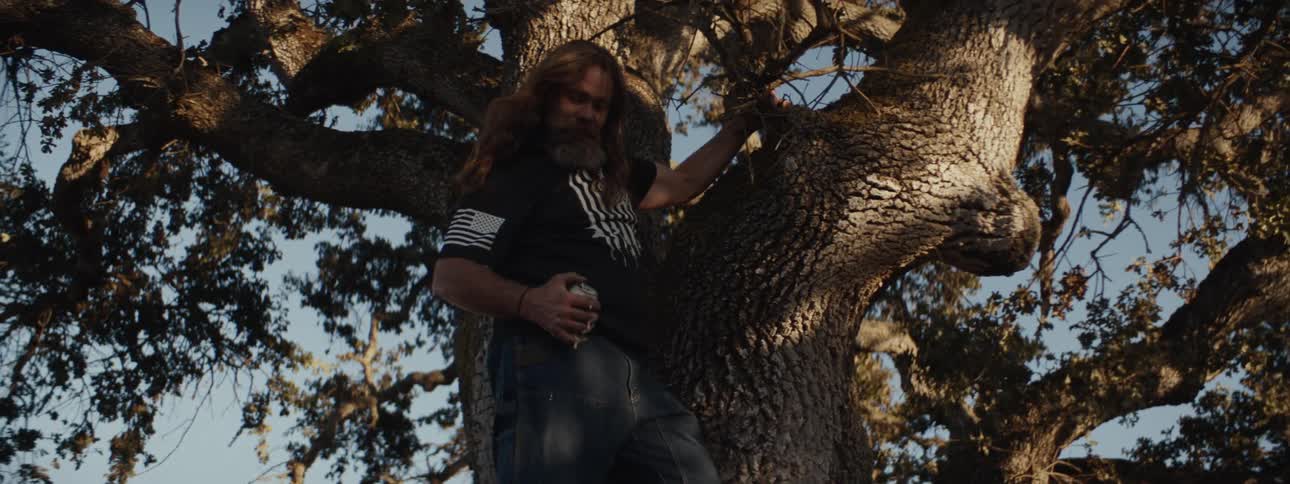 Man handing a can of beer to another man who is on a tree trunk