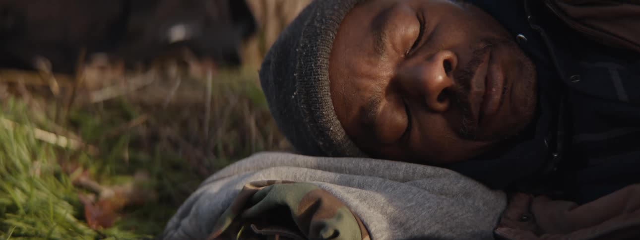 A man sleeps in a makeshift camp near a tree in the grass in the morning