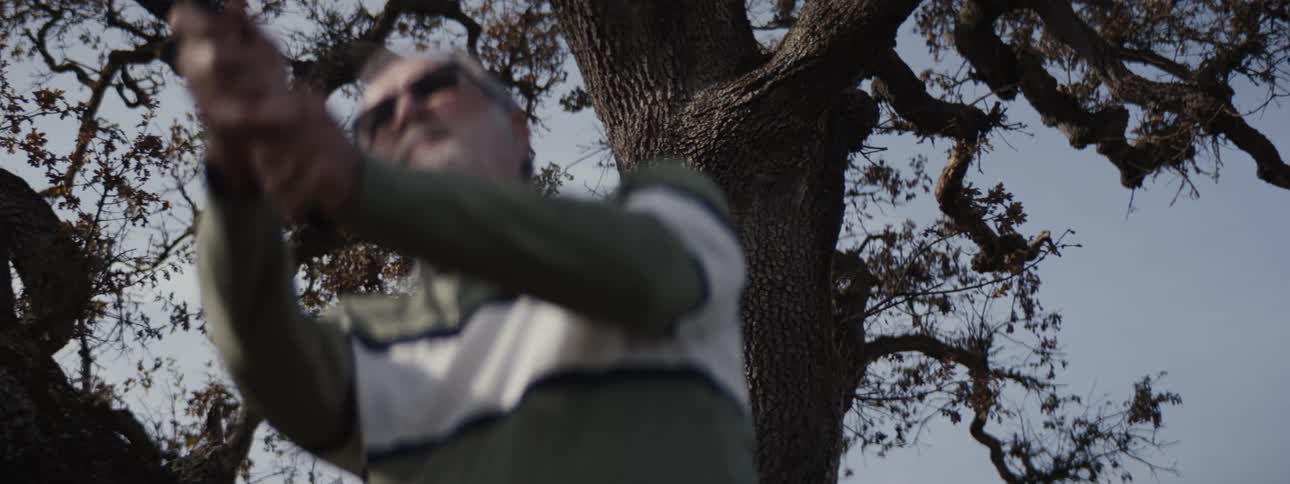 An older man swings a golf club while standing under a large tree