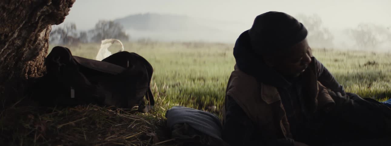 A man sleeping on a sleeping bag in a mist covered grassy field wakes up in the morning