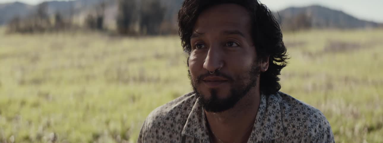 A closeup of a young bearded man listening attentively to someone else sitting on a field of green grass near the mountains
