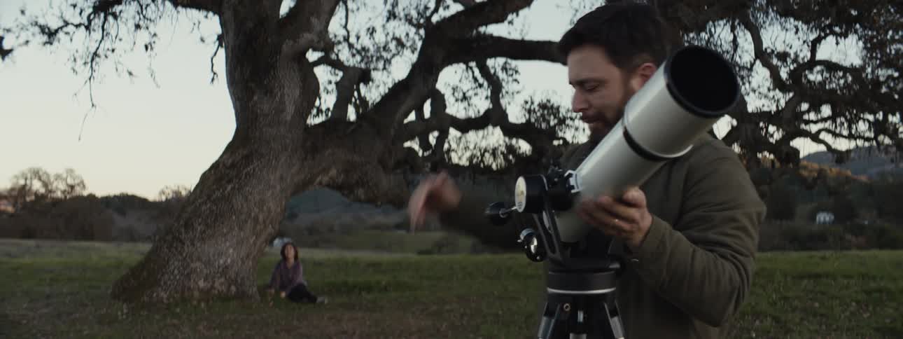 A serene outdoor scene at dusk where a bearded man in a dark jacket is adjusting a telescope while a young girl sits under a large sprawling tree in the background gazing towards him