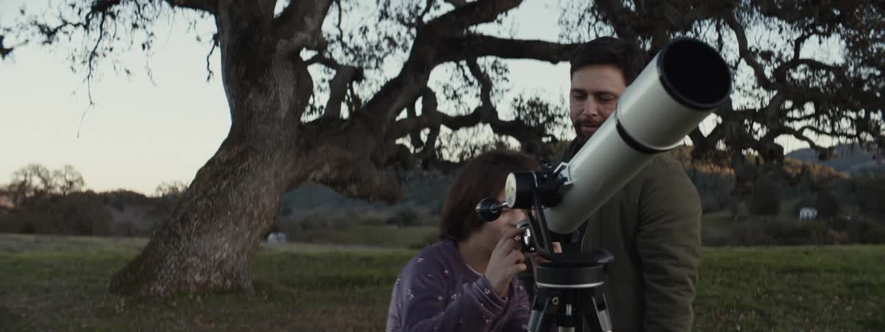 A young girl in a purple sweater watches intently as a man adjusts a large telescope in an open field with a sprawling tree and rolling hills in the background