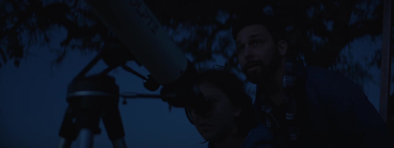 A young bearded man teaching a girl how to use a telescope outdoors at night
