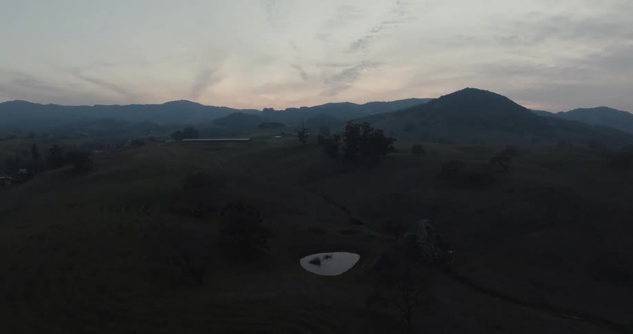 A vast shadowy landscape of rolling hills and distant mountains stretches across the frame under a dusky sky with a small bright reflective surface of water glistening in the lower portion of the scene