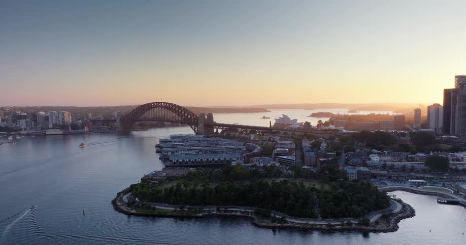 A drone shot of the sun setting on sydney harbour the harbour bridge is central the opera house is in behind it