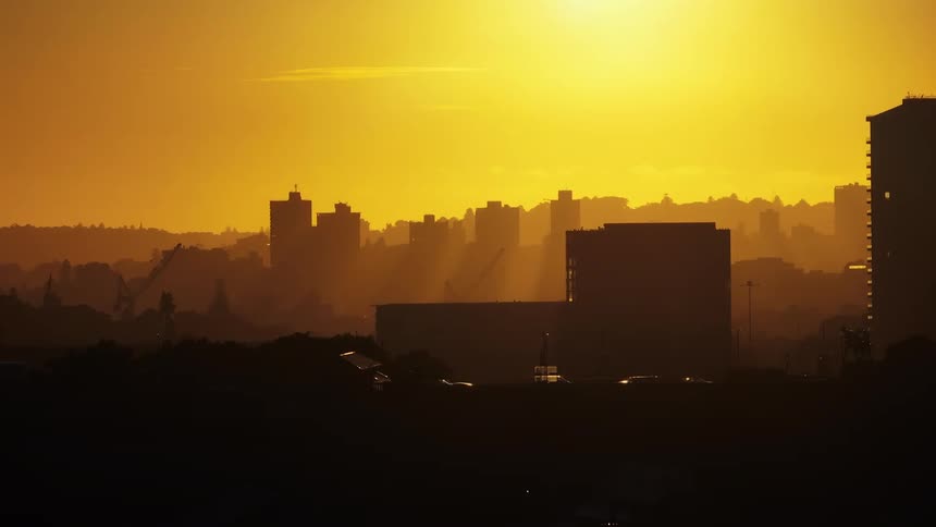 Silhouetted cityscape against a bright hazy yellow sky with a sun flare glowering over distant structures