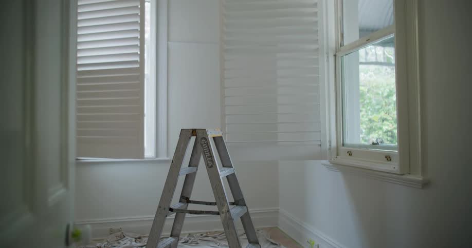 A ladder stands in a room with white walls and closed window blinds