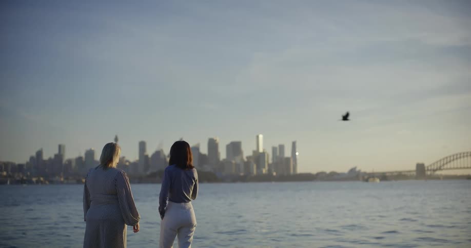 Two women side by side facing the sea and a metropolis with skyscrapers in the background on the horizon