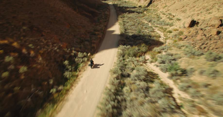 A lone motorcycle rider travels down a dusty road through a canyon with rocky cliffs on either side