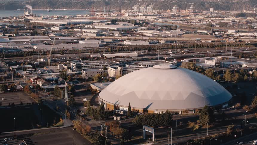 Aerial view of the tacoma domes distinctive domed roof in the foreground with a sprawling cityscape and mount rainiers peak faintly visible in the background under a mostly sunny sky