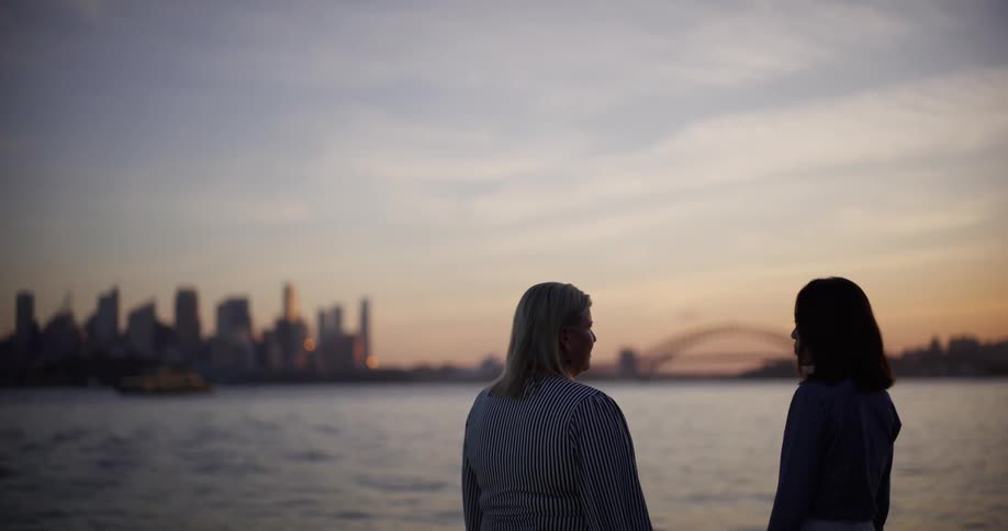 Two women talking by the sea with city buildings in the background