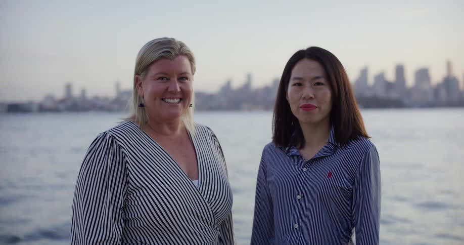 Two women standing near a body of water with a city skyline in the background one is smiling at the camera while the other has her eyes closed and lips slightly pursed