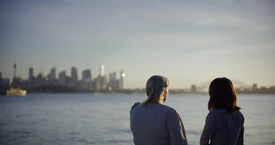 Two women wearing striped shirts are standing in front of a waterfront with a city skyline in the background