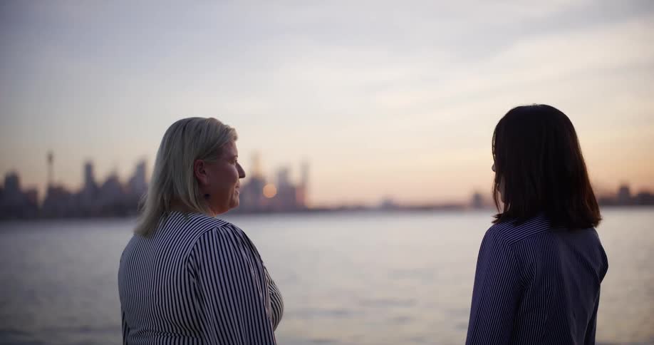Two women stand by the seaside with a city skyline in the background at dusk