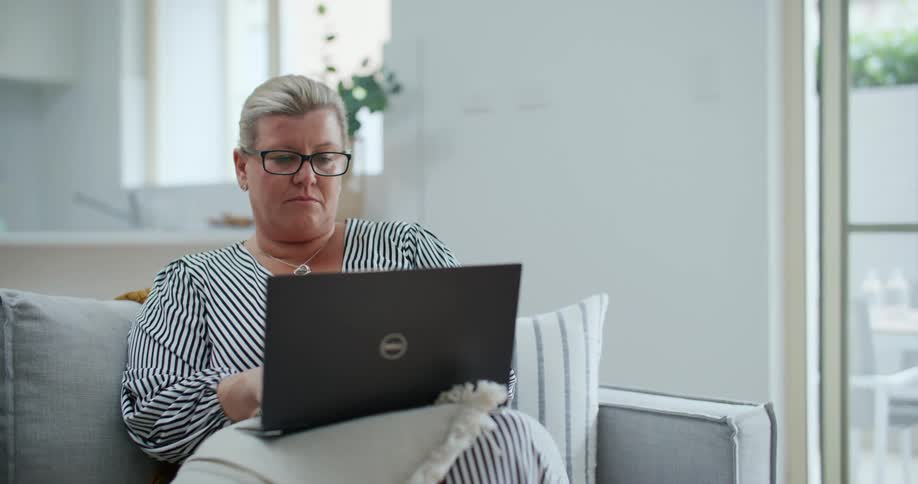 A woman with white hair and wearing glasses is sitting on a sofa and using a laptop