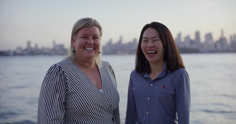 Two women standing smiling as they look forward and behind them there is a sea and a city behind