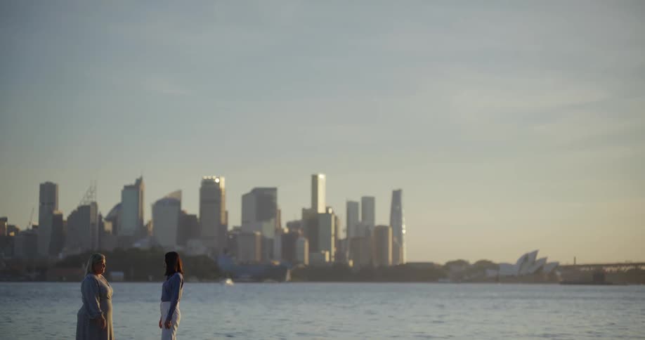 Two women talking face to face by the sea with a city in the background
