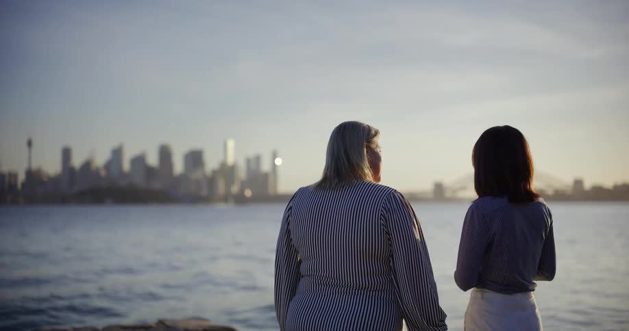 Two women with their backs turned are looking at the sea where there is a city in the distance