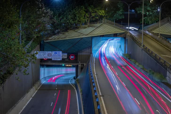A multi level highway interchange at night illuminated by streetlights and the red streaks of taillights with a building facade and trees visible on the side