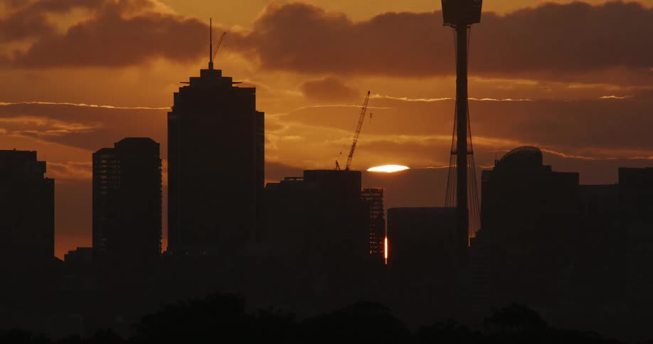City buildings are silhouetted against a bright cloudy sky with a sunset featuring a tall tower and construction cranes