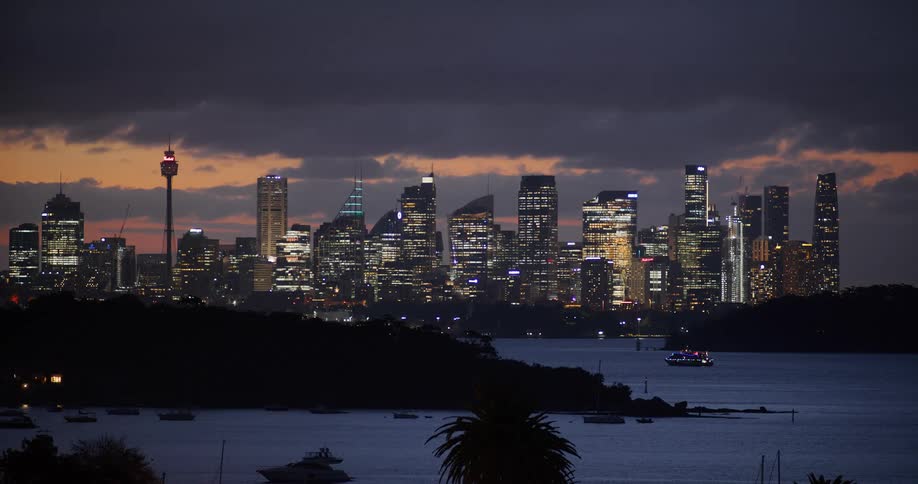Night view of an illuminated skyline with tall buildings reflecting on the water under an orangehued sky and dark clouds