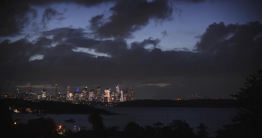 A dimly lit cityscape skyline glows with warm lights under a cloudy dusk sky reflected in the dark water of a harbor