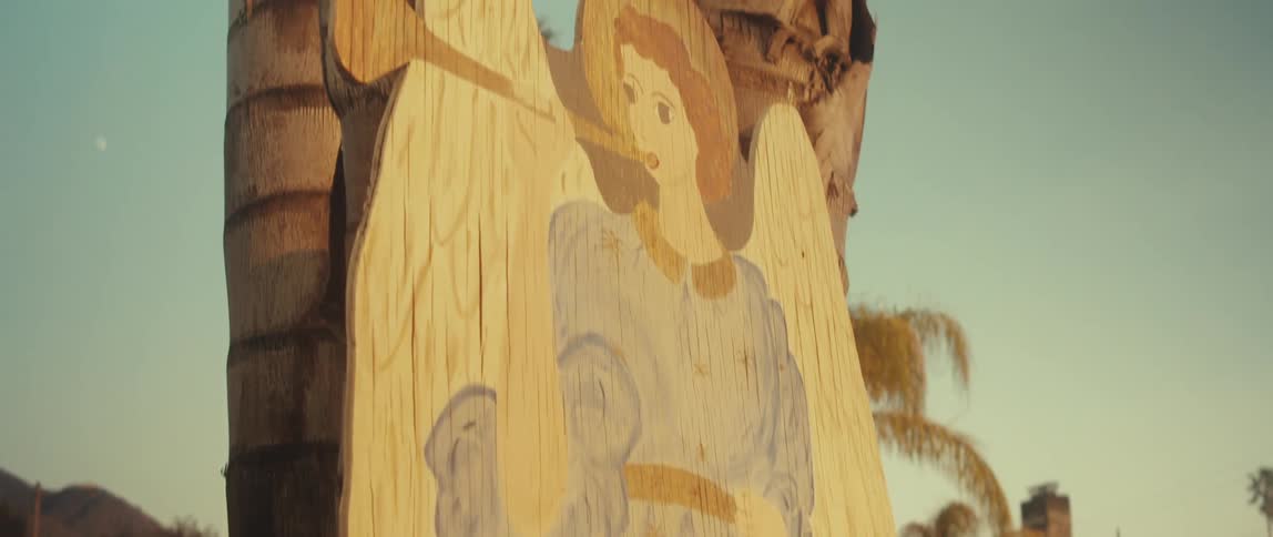 A closeup shot shows a wooden cutout of an angel playing a trumpet attached to two palm tree trunks against a light blue sky