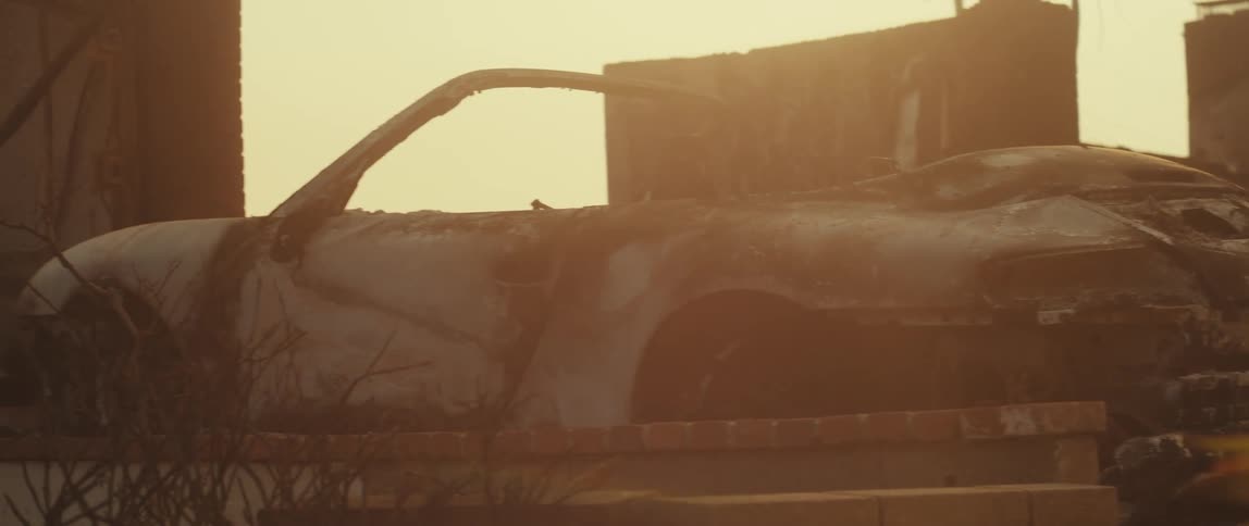 A rusted abandoned car sits in a desolate area with a dilapidated building in the background under a hazy sky