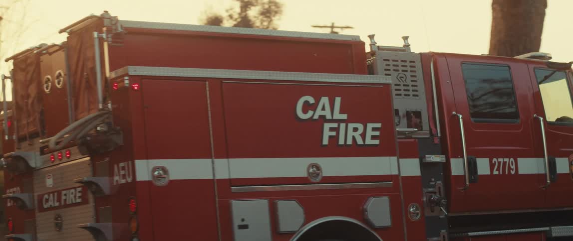 A red fire truck is driving near a destroyed building with a visible car wrecks in the background
