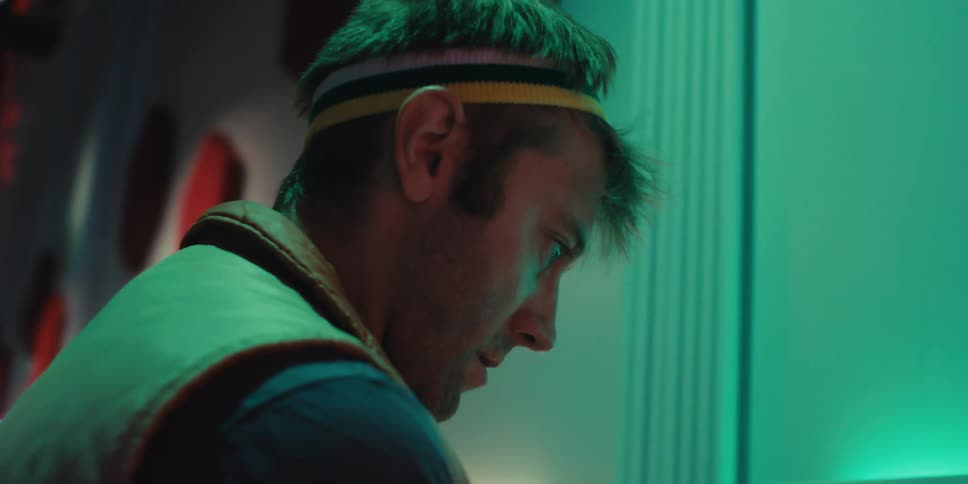 A man with a headband is playing an arcade game and the screen is lighting up his face