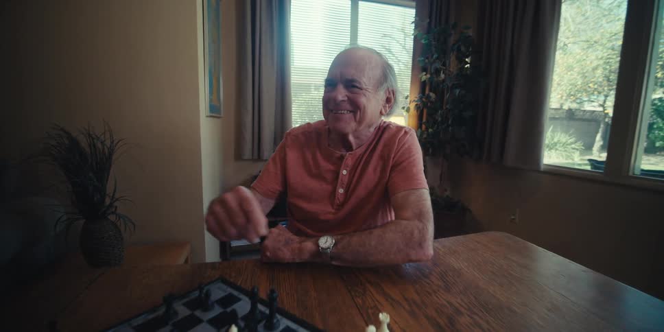 Elderly man laughs while playing chess at home with a younger man