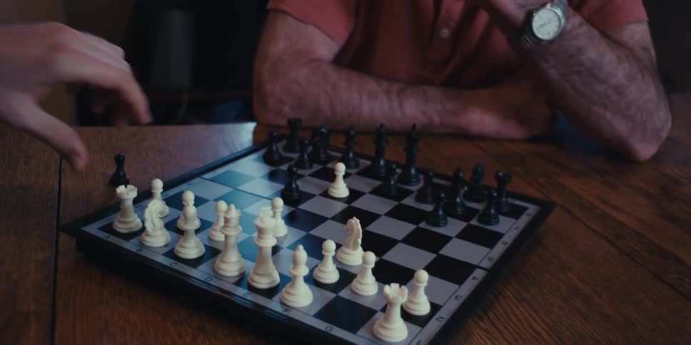 An elderly man concentrates on a chess game at home with a friend or family member