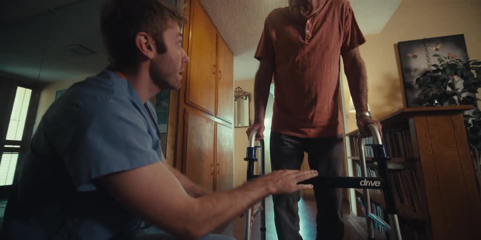 Nurse crouching down helping elderly man walk with walker down hallway