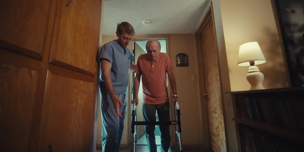 A male nurse assists an elderly patient with walking using a walker at home