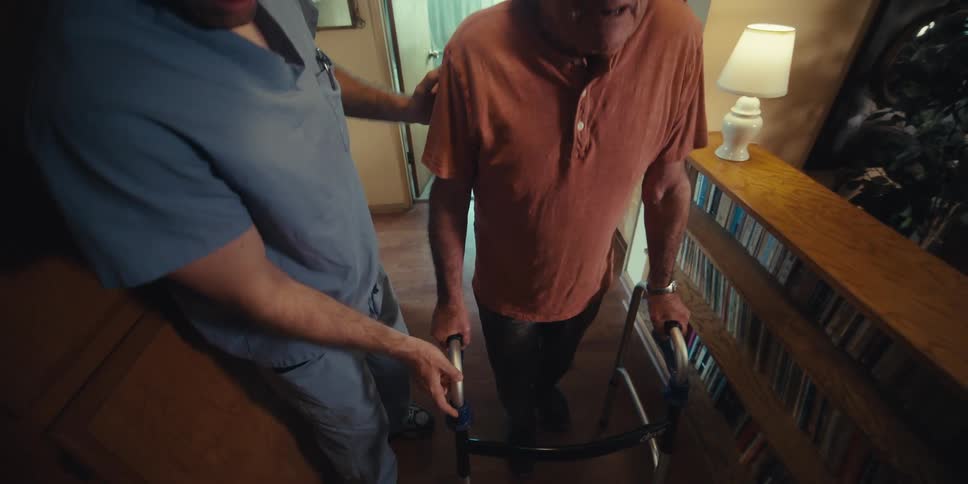 Elderly man walking down a hallway with a walker and a nurse by his side