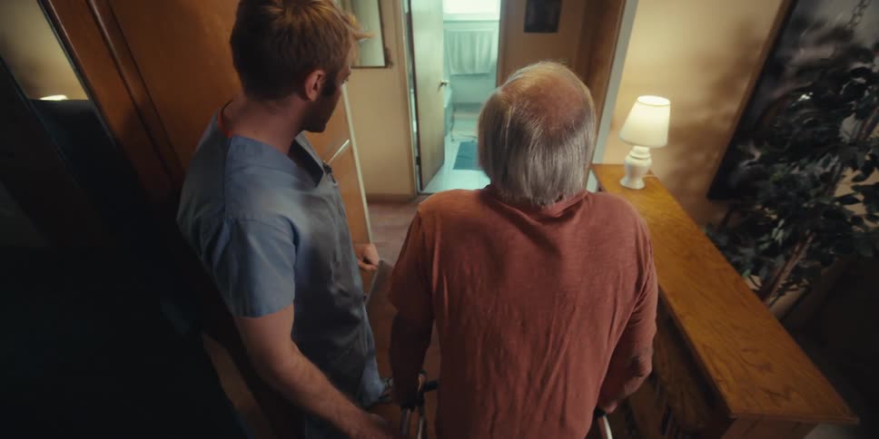 A man is walking through a door with a walker being guided by a nurse