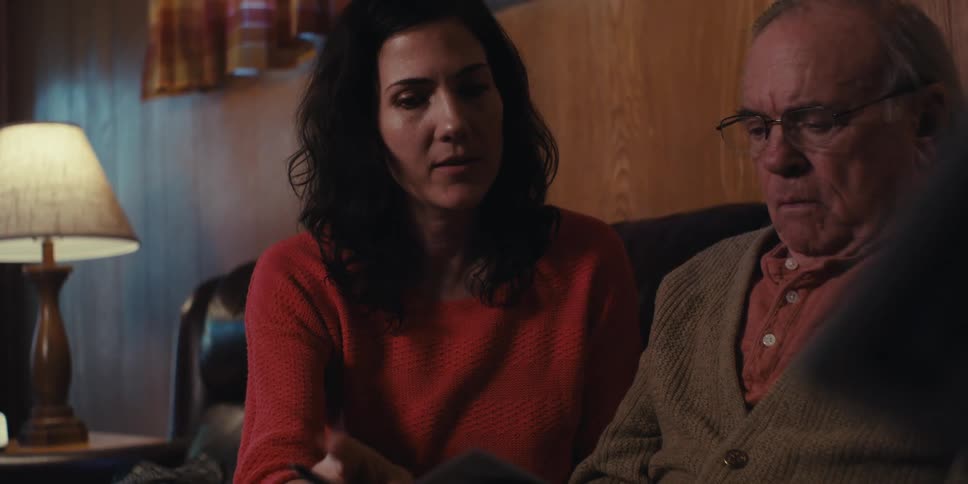A person pointing a pen at a piece of paper held by an elderly man who is sitting next to a woman in a red shirt