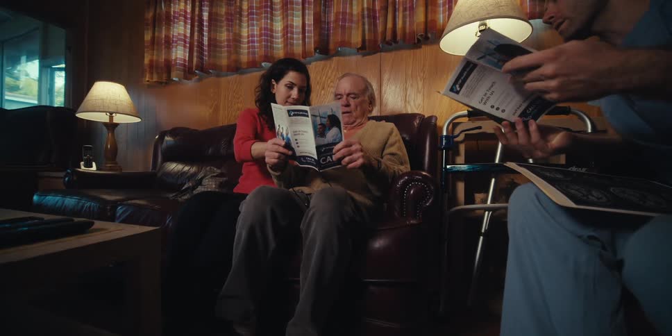 Nurse presenting a brochure to an elderly gentleman and his adult daughter who are sitting next to him on a living room sofa