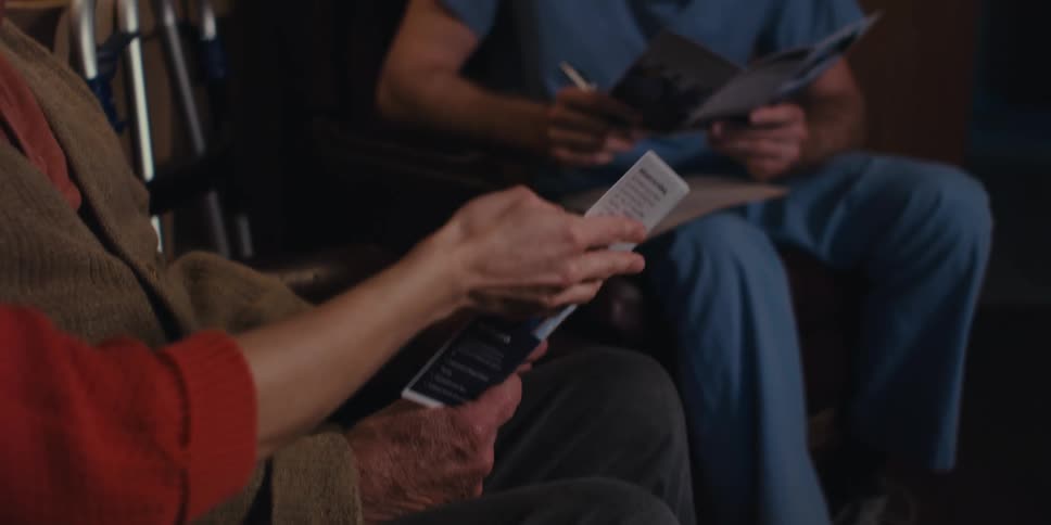 An elderly persons hand holding a paper and a man in blue clothes is sitting in front and looking