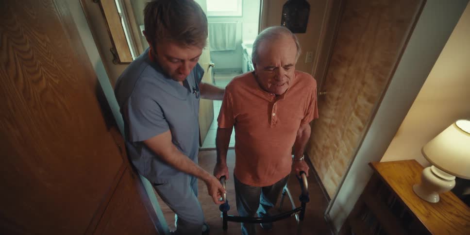 Nurse helping an elderly man walk with a walker down the hallway of his home