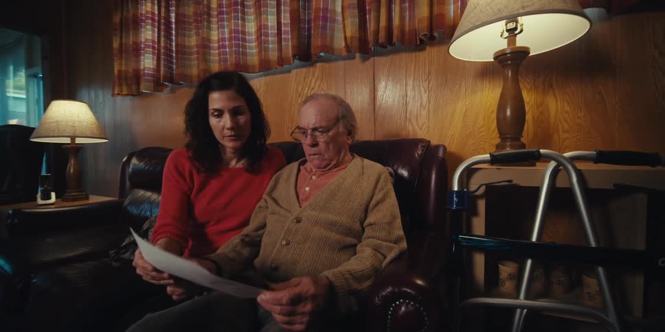 A man pointing to a paper with photos that is in the hand of an elderly man who is sitting on the sofa next to a woman